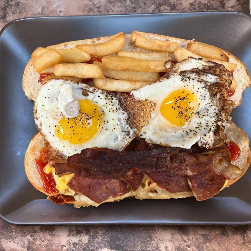 A Cedar Grill & Pizza breakfast sandwich with fried eggs, bacon, and fries on a dark plate.